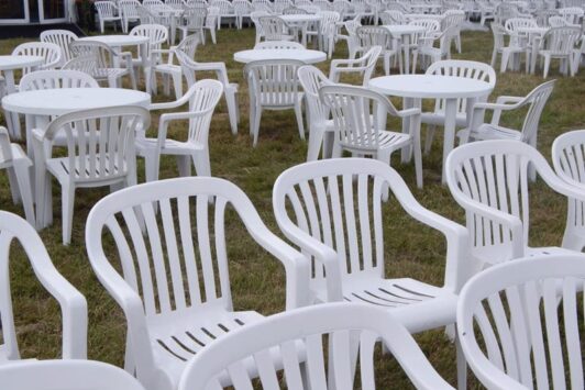 Rows of white plastic tables and chairs set up outdoors on grass.