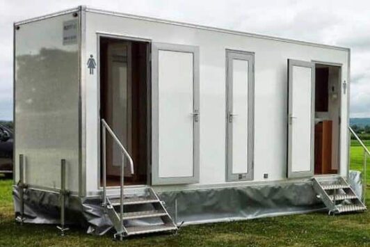 Portable toilet trailer with multiple doors and steps, set up outdoors on grass.