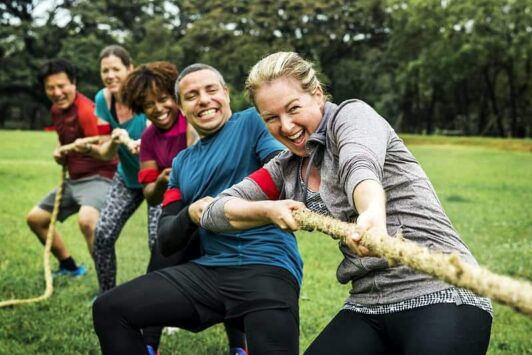 Smiling adults playing tug of war outdoors on a grassy field.