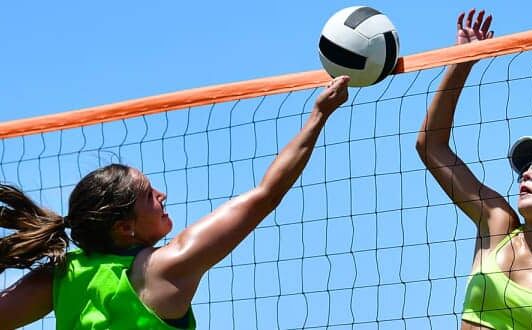 Two women play beach volleyball, reaching above the net for the ball under a clear blue sky.