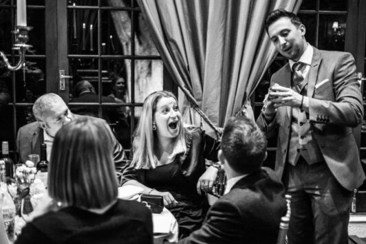A magician performs a trick at a table, surprising a group of people in formal attire.