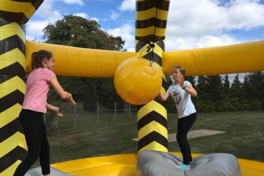 Two girls play with a large yellow inflatable wrecking ball outdoors on a bouncy structure.